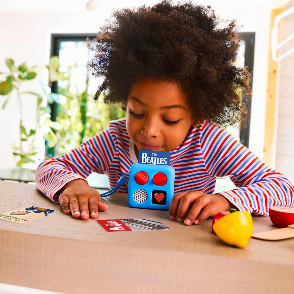 Child at a table with a blue Yoto Mini, orange dials, a pixel heart on the display, a Beatles card inserted and spare cards beside wooden play food