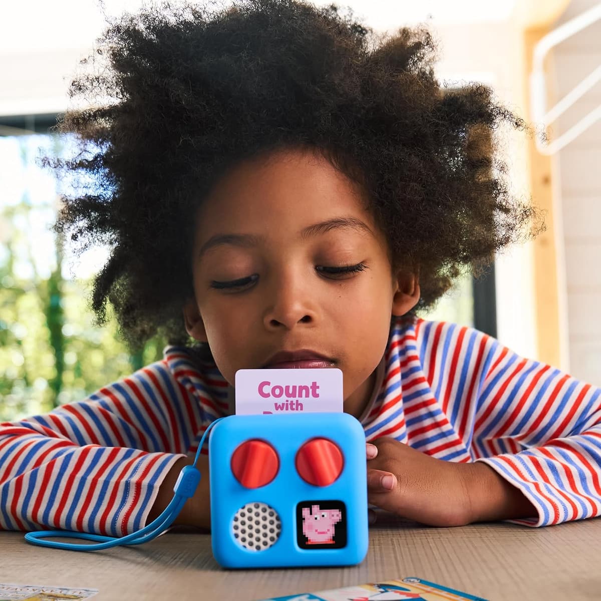 Young child at a wooden table looking at a light-blue Yoto Mini with orange dials, a pixel character on the display and a counting card in the top slot